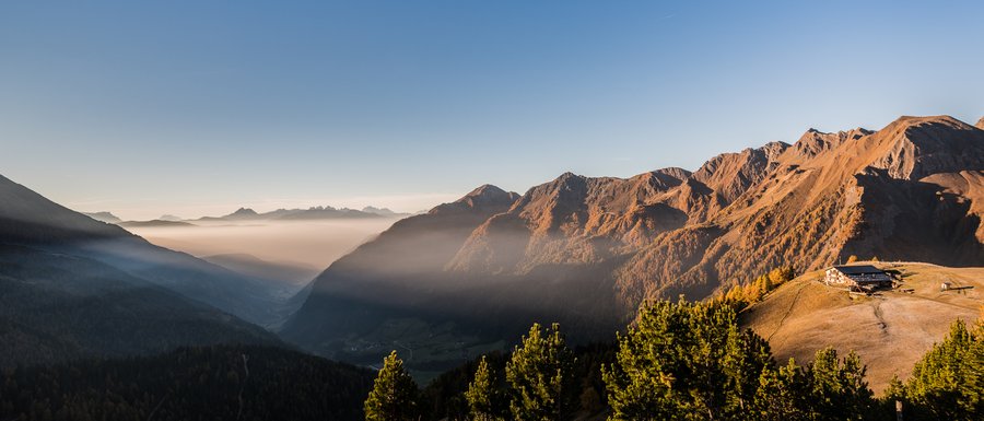 Find your accommodation in South Tyrol The picture shows a picturesque mountain landscape at sunrise or sunset, with deep valleys covered in fog and sunlit mountain slopes glowing in warm orange tones. On a ridge to the right in the image stands a mountain hut, surrounded by grassy hills and coniferous trees.