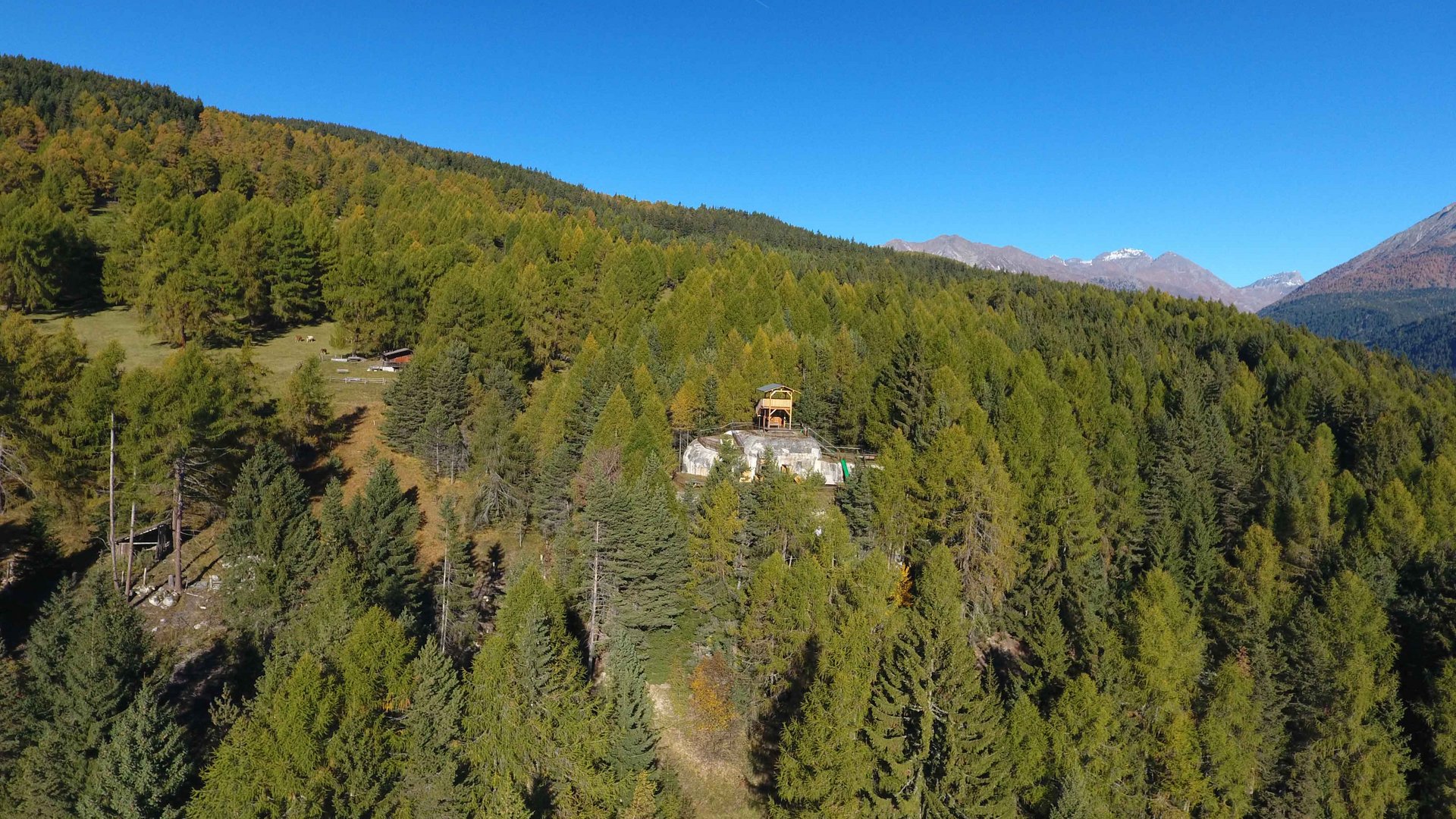 Het recreatiegebied in Spinges/Zuid-Tirol De afbeelding toont een klein gebouw dat zich op een beboste heuvel bevindt, omgeven door dicht bos en bergen op de achtergrond. De lucht is helder en blauw, en het terrein is bedekt met veel groene bomen.