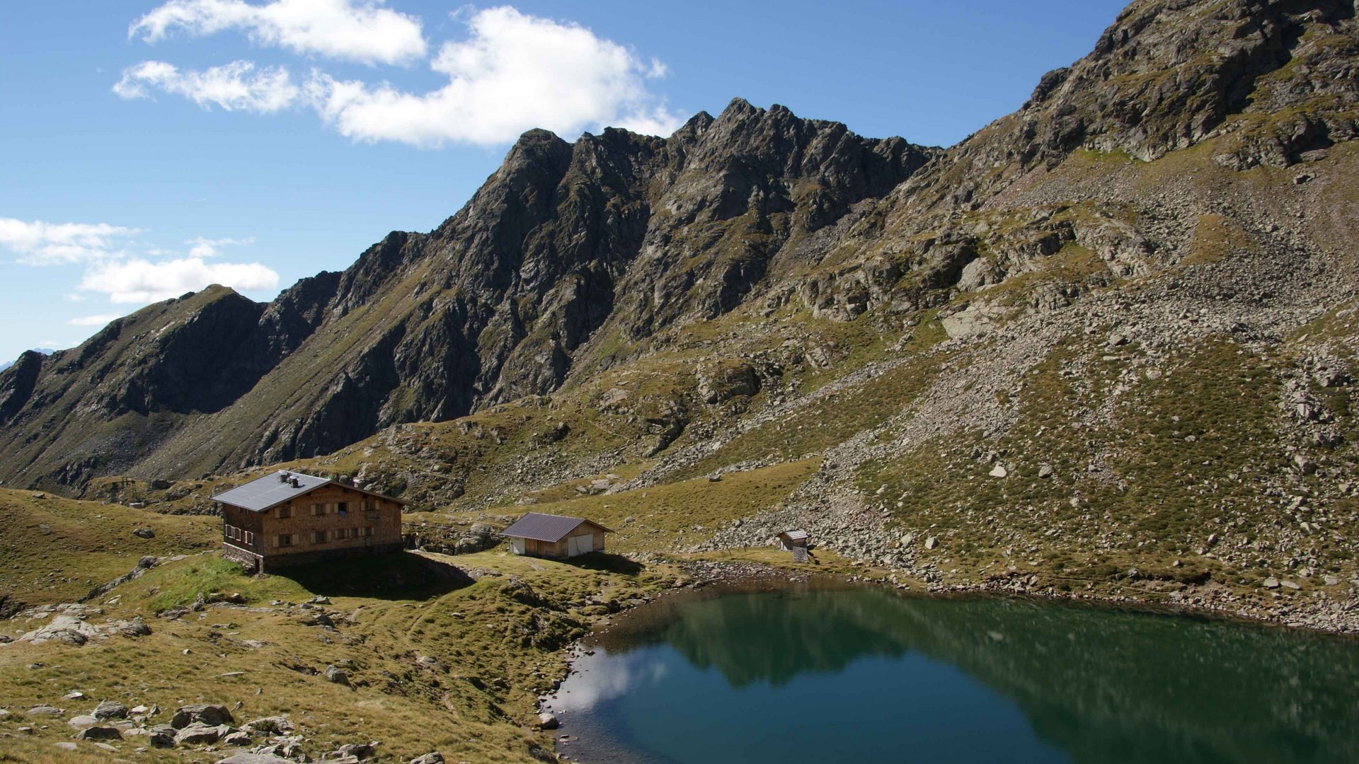 The Pfunders High Mountain trail The picture shows a mountain hut next to a calm, clear mountain lake in a rocky alpine landscape. Steep, rugged mountains rise in the background under a blue sky with scattered clouds.