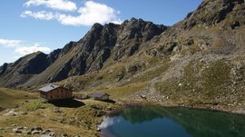 The Pfunders High Mountain trail The picture shows a mountain hut next to a calm, clear mountain lake in a rocky alpine landscape. Steep, rugged mountains rise in the background under a blue sky with scattered clouds.