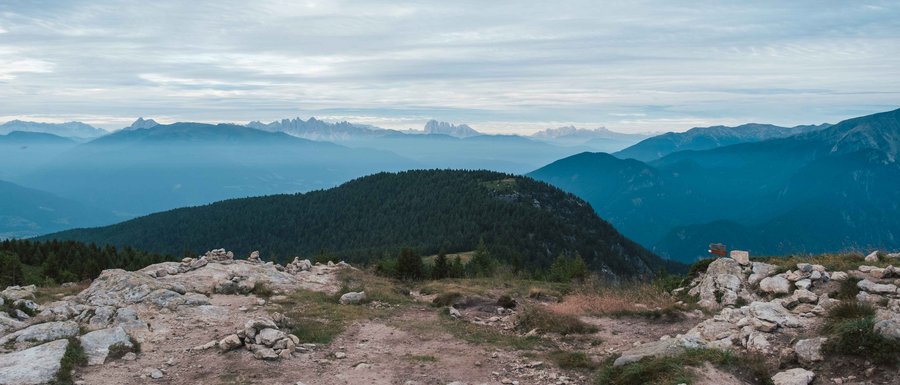 Find your accommodation in South Tyrol The picture shows a mountain landscape with several layers of hills and mountains stretching into the distance. The foreground consists of a rocky terrain, while in the background wooded hills and more distant mountain ranges can be seen under a cloudy sky.