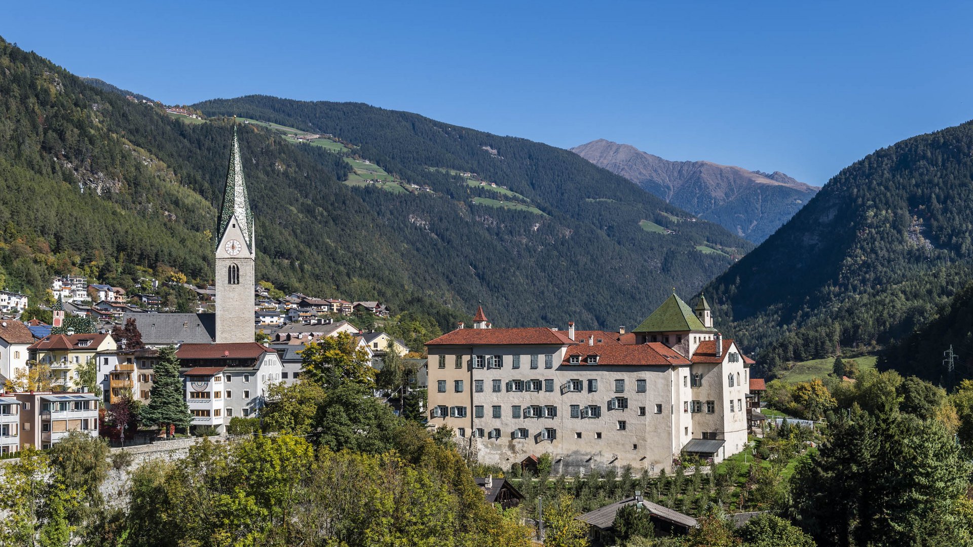 Mühlbach in the Pustertal valley The picture shows a picturesque small town in a valley, surrounded by forested hills and mountains. In the center stands a church with a tall, pointed tower, and next to it is a large building with red roofs and many windows.