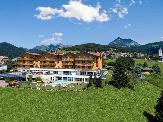 Willkommen in der Ferienregion Gitschberg Jochtal! Das Bild zeigt ein großes, modernes Hotelgebäude in einer alpinen Umgebung. Im Vordergrund liegt eine grüne Wiese, während im Hintergrund Berge und eine kleine Stadt zu sehen sind.