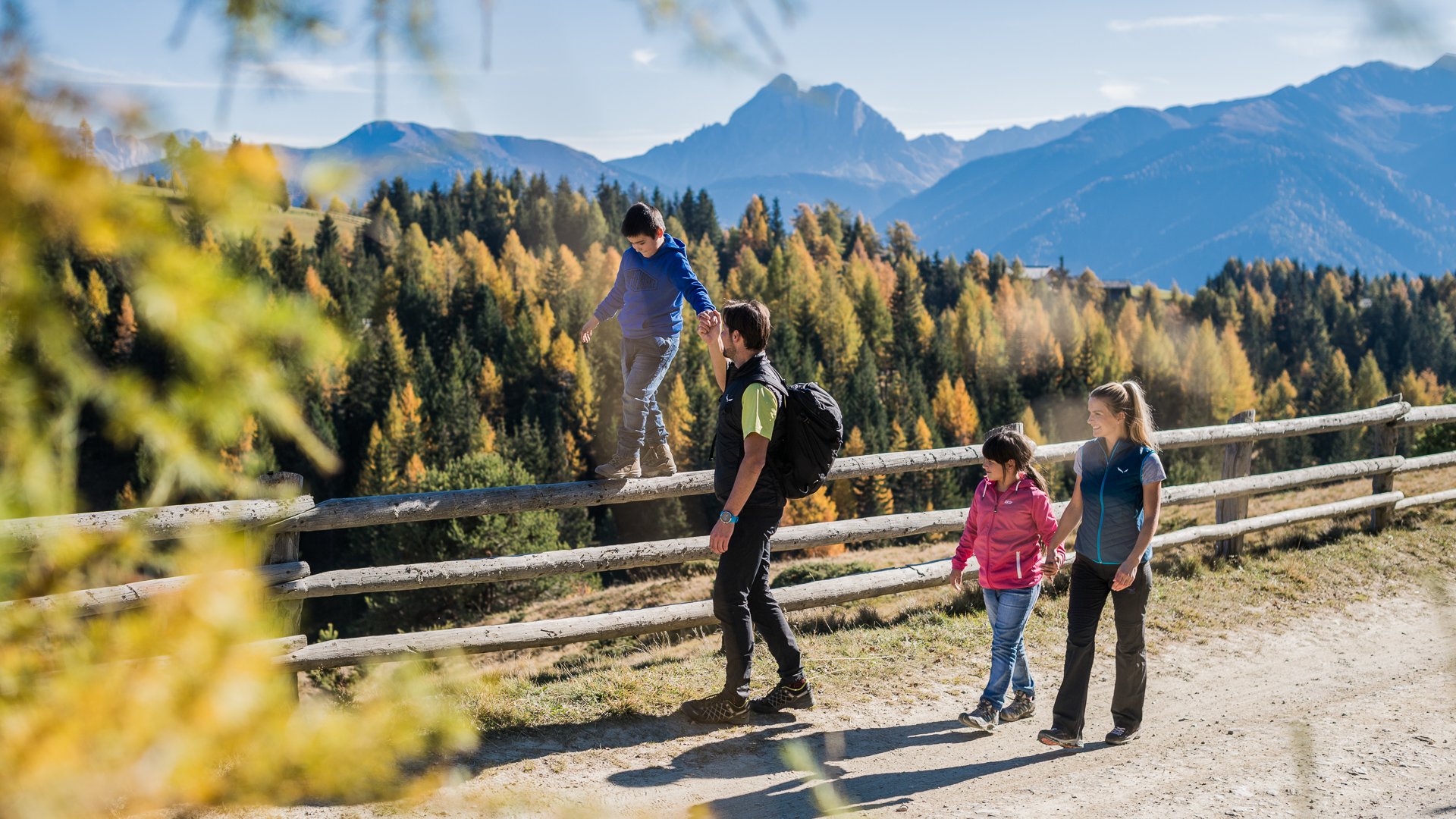 De Rodenecker-Lüsner Alm De afbeelding toont een gezin dat wandelt op een pad in de bergen. Een kind balanceert op een houten hek terwijl de vader zijn hand vasthoudt, en op de achtergrond zijn kleurrijke herfstbomen en bergen te zien.
