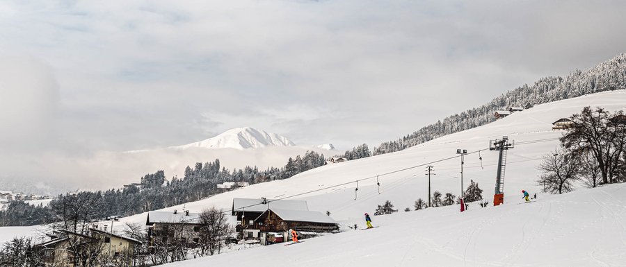 Find your accommodation in South Tyrol The picture shows a snow-covered landscape with a ski lift and several skiers on a slope. In the background, snowy hills and mountains can be seen, partially obscured by clouds.