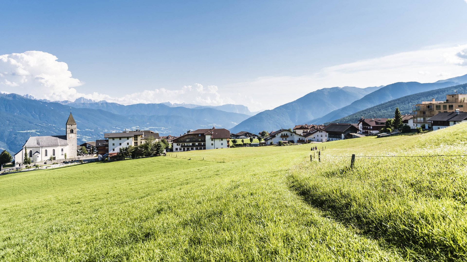 Horská vesnice Meransen Obrázek ukazuje malebnou vesnici s kostelem vlevo, obklopenou zelenými loukami a roztroušenými domy. V pozadí se rozprostírají zalesněné hory pod jasně modrou oblohou s několika mraky.