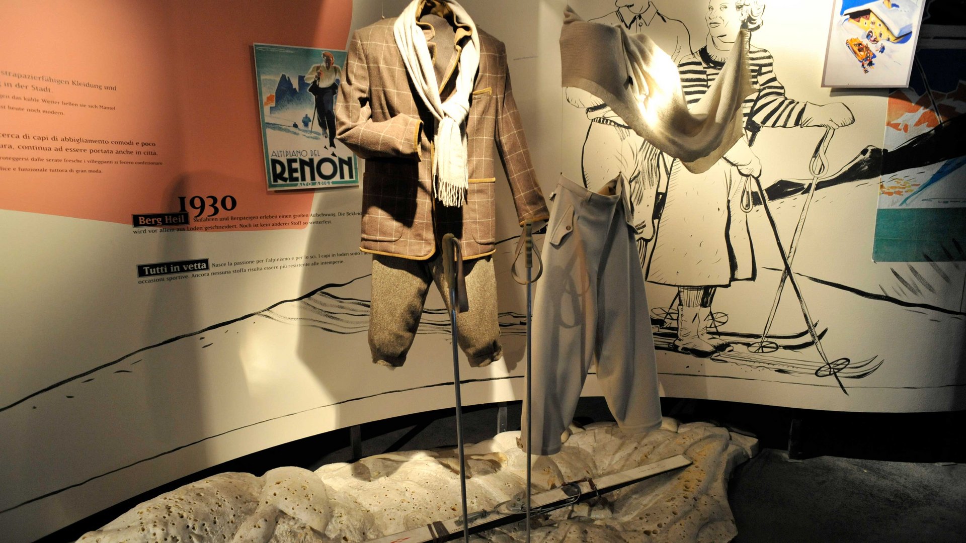 Zážitkový svět lodenu ve Vintlu/Pustertal Obrázek ukazuje výstavu lyžařského oblečení z 30. let 20. století, která je prezentována na výkladních figurínách. V pozadí jsou ilustrace lyžařů a starý plakát, které zobrazují módu a lyžařský sport té doby. Pár lyží leží na zemi před oblečením.