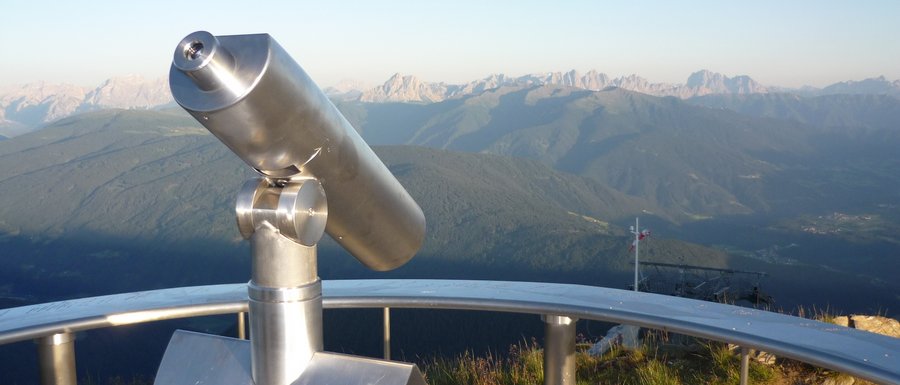 Find your accommodation in South Tyrol The image shows a permanently installed binocular at a viewpoint, directed towards a vast mountain landscape. In the background, mountain ranges and valleys can be seen under a clear blue sky.