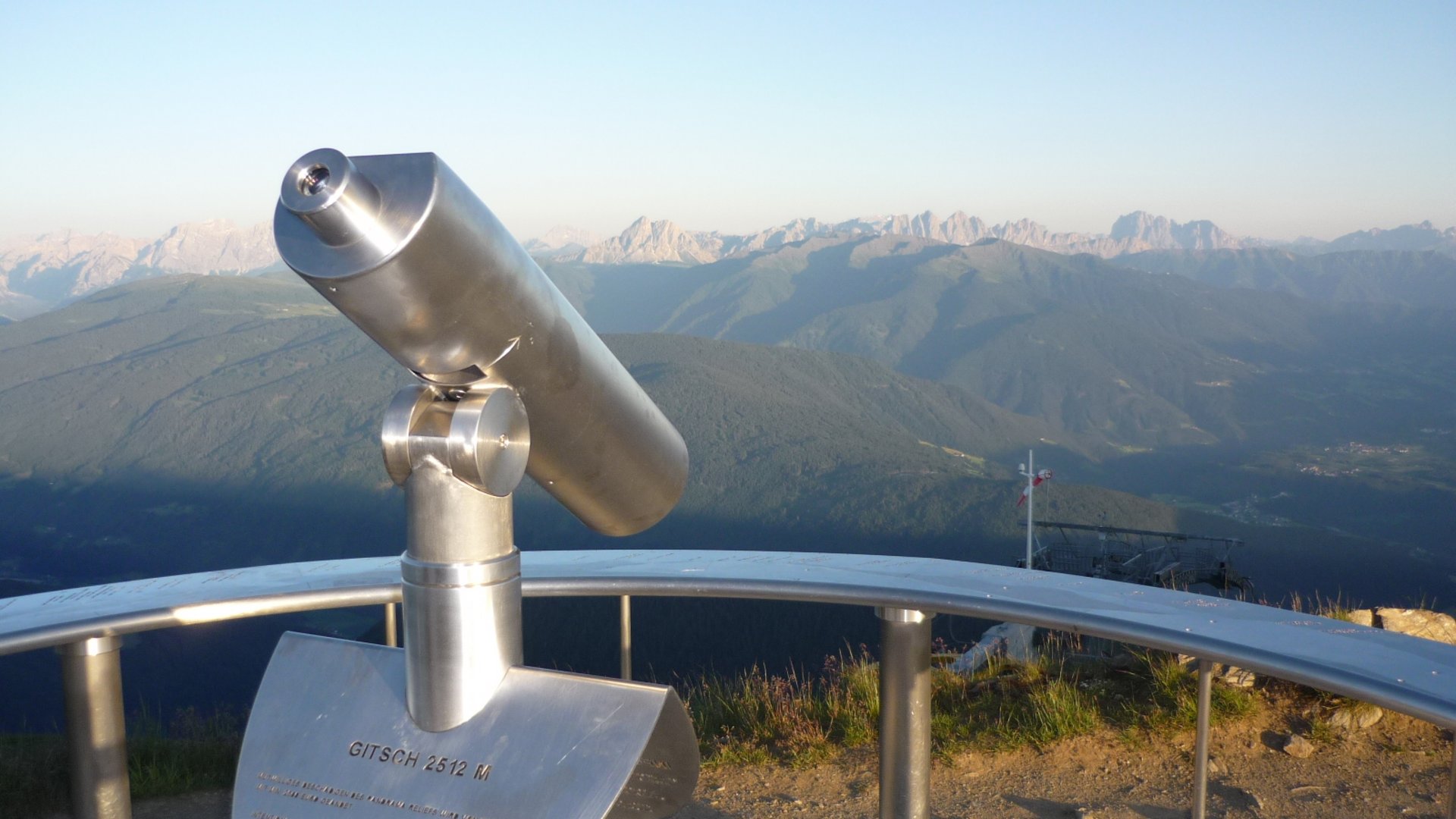 The Gitschberg panoramic platform in Meransen, Italy The image shows a permanently installed binocular at a viewpoint, directed towards a vast mountain landscape. In the background, mountain ranges and valleys can be seen under a clear blue sky.