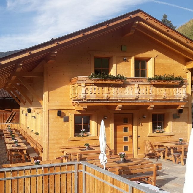 Hofschänke Lechnerhof Eine Berghütte aus hellem Holz mit einem Balkon und Blumenkästen steht an einem Berghang. Vor der Hütte sind mehrere Holztische und Bänke mit Sonnenschirmen auf einer Terrasse aufgestellt.