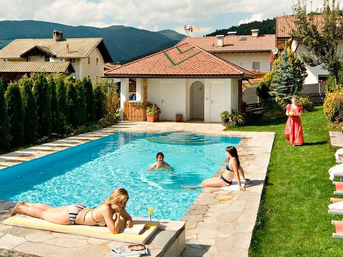 Willkommen in der Ferienregion Gitschberg Jochtal! Das Bild zeigt einen Außenpool in einem gepflegten Gartenbereich, umgeben von Sonnenliegen. Zwei Frauen sonnen sich am Pool, während eine Person im Wasser schwimmt. Im Hintergrund sind mehrere Gebäude und Berge zu sehen, die eine ruhige und idyllische Umgebung schaffen.
