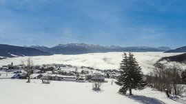 Das Bergdorf Meransen Das Bild zeigt eine schneebedeckte Landschaft mit einer kleinen Ortschaft im Vordergrund, während sich im Hintergrund ein Meer aus Wolken erstreckt. Darüber erheben sich majestätische, schneebedeckte Berge unter einem klaren, blauen Himmel.