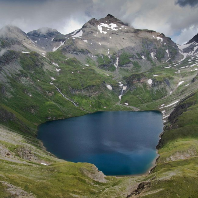 Pěší turistika v Jochtalu a Gitschbergu a další Obrázek zobrazuje vysoko položené horské jezero obklopené strmými zelenými horskými svahy a zasněženými vrcholy. Obloha je zatažená, což scéně dodává dramatickou atmosféru.