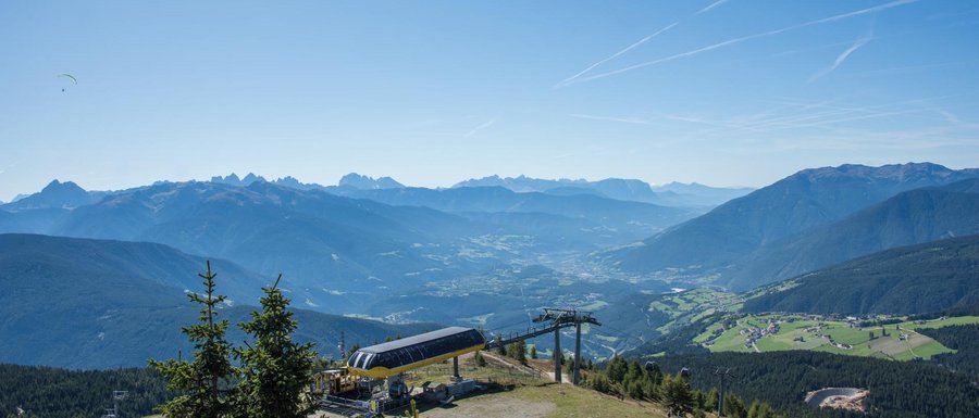 Find your accommodation in South Tyrol The picture shows a vast mountain landscape under a clear blue sky. In the foreground, there is a cable car station on a mountain peak, and in the background, several mountain ranges as well as a valley with scattered villages and forests can be seen. Two paragliders are flying high in the sky above the scene.