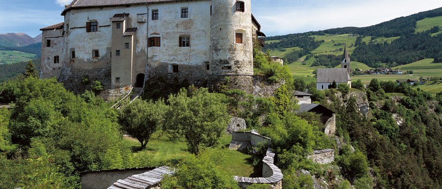 Find your accommodation in South Tyrol The picture shows an old castle on a green hill, surrounded by trees and a stone wall. In the background, there is a church and a rural landscape.