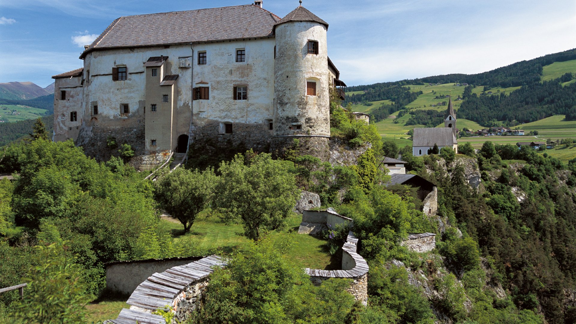 A mighty landmark: Castle Rodenegg in Rodeneck The picture shows an old castle on a green hill, surrounded by trees and a stone wall. In the background, there is a church and a rural landscape.