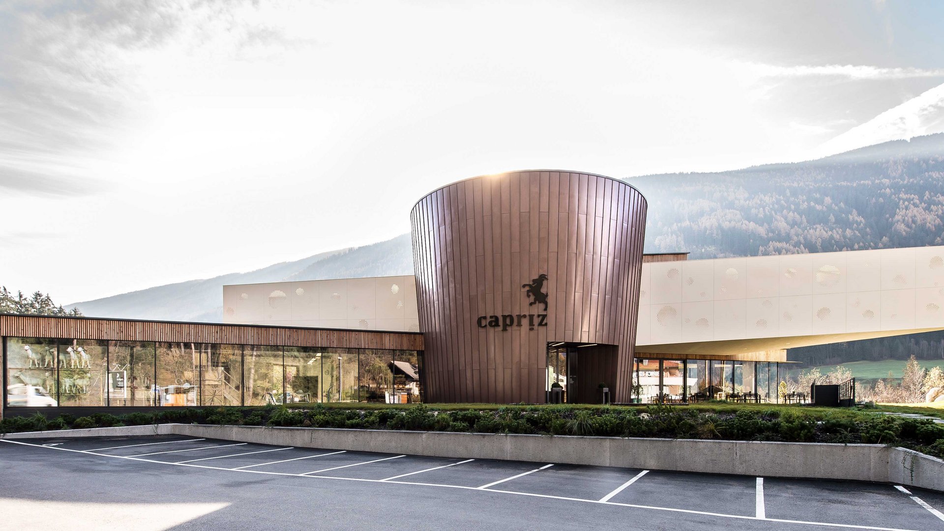 De Feinkäserei Capriz De afbeelding toont een modern gebouw met een ronde koperkleurige gevel en grote glasramen. Op het gebouw staat de tekst "Capriz" en ervoor is een lege parkeerplaats, terwijl op de achtergrond beboste heuvels en bergen te zien zijn.