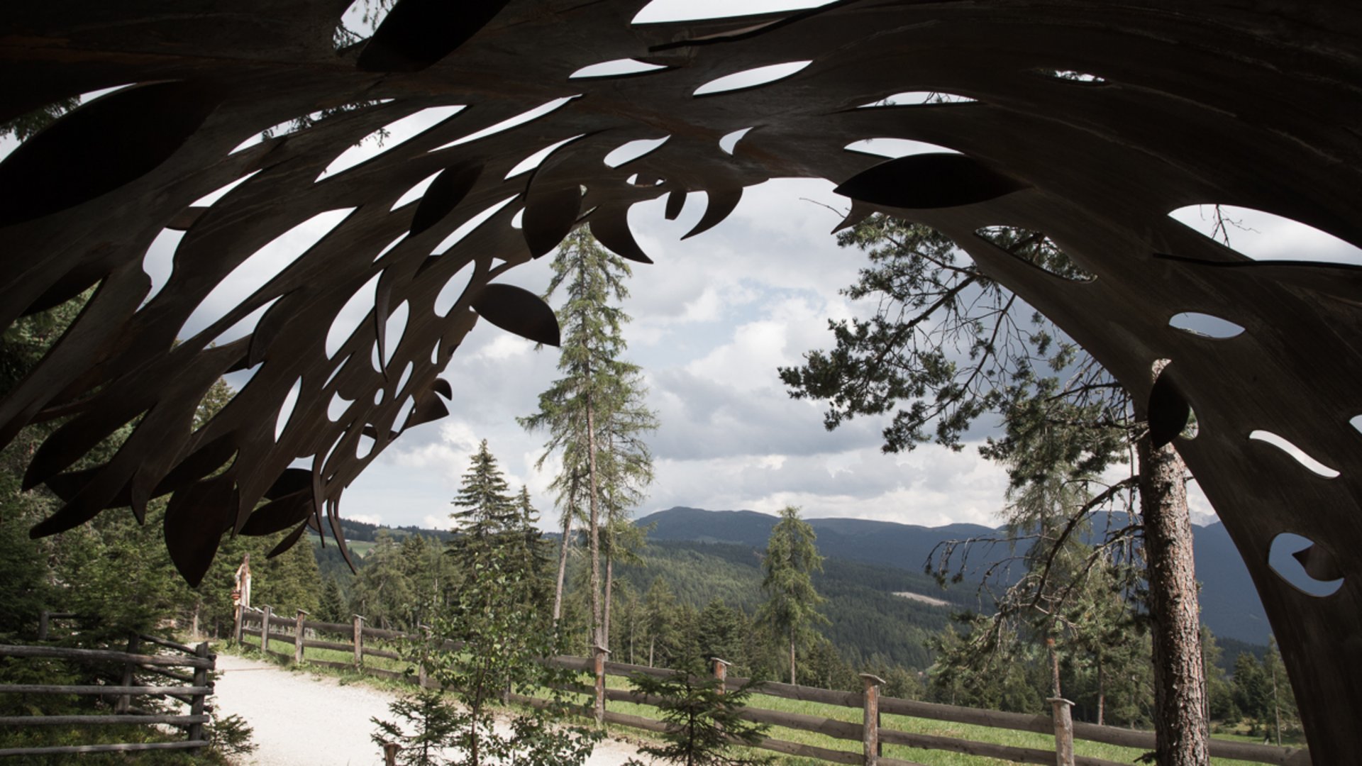 The Creation trail in Rodeneck The picture shows a view through a metallic sculpture with organic shapes. In the background, there are trees, a path with a wooden fence, and wooded hills under a cloudy sky.