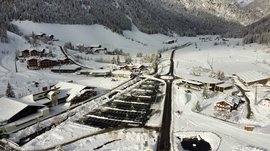 Vals: das Almendorf Das Bild zeigt ein verschneites Dorf aus der Vogelperspektive. Die Straßen und Häuser sind von Schnee bedeckt, und ein Parkplatz mit geräumten Stellplätzen ist im Vordergrund zu sehen. Im Hintergrund erstreckt sich ein bewaldeter Berghang.