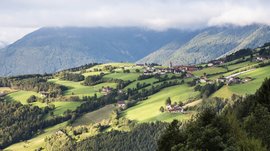 The Alpine hamlet of Spinges The picture shows a gentle, green hilly landscape with scattered houses and a small church with a red tower. In the background, forested mountain slopes stretch out, partially obscured by clouds.