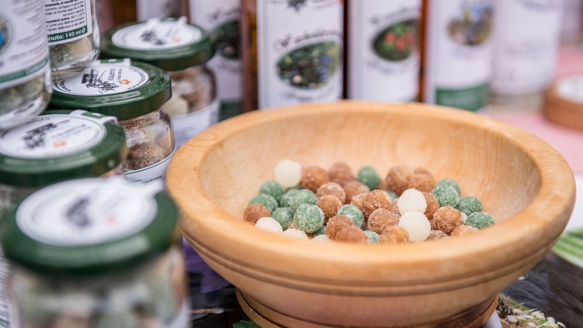 Nakupování v Mühlbachu/Itálie Obrázek ukazuje dřevěnou misku naplněnou barevnými bonbóny. V pozadí stojí několik sklenic se zelenými víčky, které jsou také naplněny bonbóny nebo podobnými sladkostmi. Vypadá to jako tržní stánek nabízející ručně vyráběné výrobky.