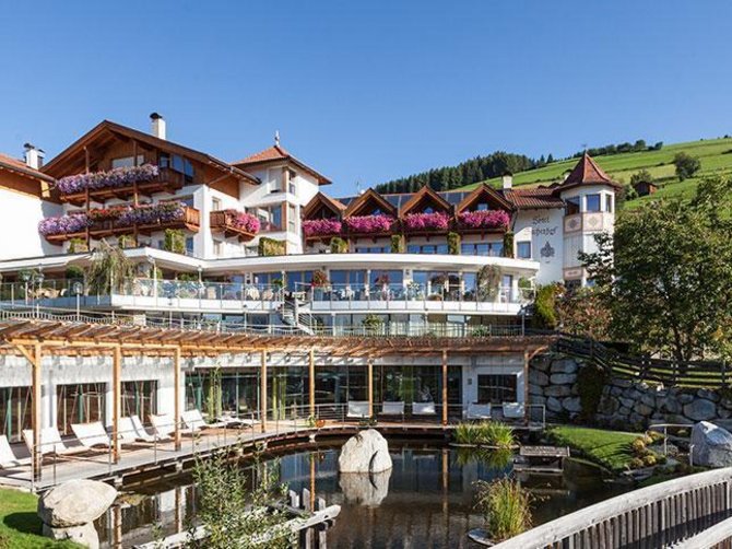 Willkommen in der Ferienregion Gitschberg Jochtal! Das Bild zeigt ein großes, elegantes Hotel mit mehreren Stockwerken und Balkonen, die mit farbenfrohen Blumen geschmückt sind. Im Vordergrund befindet sich ein kleiner Teich mit einer Brücke, und im Hintergrund erstrecken sich grüne Hügel und Wiesen.