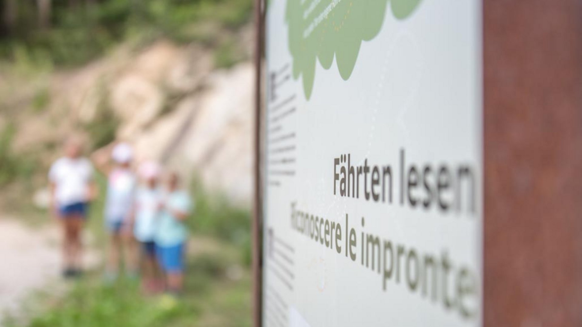 Elfí cesta (Elfenweg) u Vintlu Obrázek ukazuje ceduli s nápisem „Fährten lesen“ a italským překladem „Conoscere le impronte“. V pozadí jsou rozmazaně vidět některé děti v lesní oblasti.