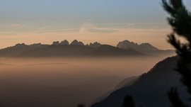 The Pfunders High Mountain trail The image depicts a mountain landscape at sunrise or sunset, where the silhouettes of the mountain peaks stand out against the orange sky. Thick layers of fog lie in the valleys, creating a mystical atmosphere.