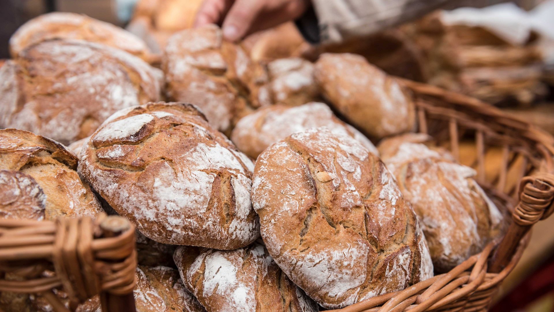 Nakupování v Mühlbachu/Itálie Obrázek ukazuje košík plný čerstvě upečených chlebů s křupavou kůrkou a lehce pomoučeným povrchem. Ruka sahá po jednom z chlebů, což naznačuje prodej nebo výběr.