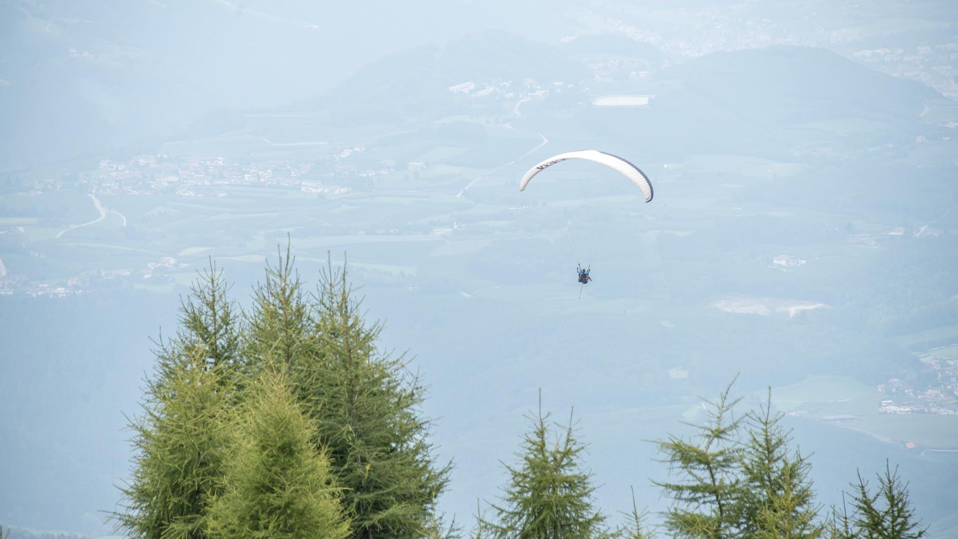 Paragliden in Zuid-Tirol De afbeelding toont een paraglider die hoog in de lucht boven een beboste berg zweeft. Op de achtergrond zijn zacht glooiende heuvels en een vallei met verspreide dorpjes en velden te zien.