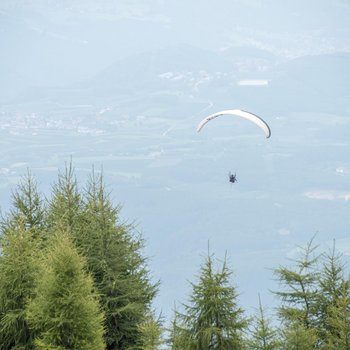 Willkommen in der Ferienregion Gitschberg Jochtal! Das Bild zeigt einen Paragleiter, der hoch in der Luft über einem bewaldeten Berg schwebt. Im Hintergrund sind sanfte Hügel und ein Tal mit verstreuten Dörfern und Feldern zu sehen.