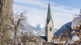 Mühlbach in the Pustertal valley The picture shows a church with a tall green church tower in a snowy village. In the background, snow-covered mountains and a blue sky are visible, while in the foreground bare trees and bushes are covered with snow.