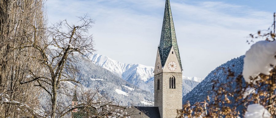 Find your accommodation in South Tyrol The picture shows a church with a tall green church tower in a snowy village. In the background, snow-covered mountains and a blue sky are visible, while in the foreground bare trees and bushes are covered with snow.