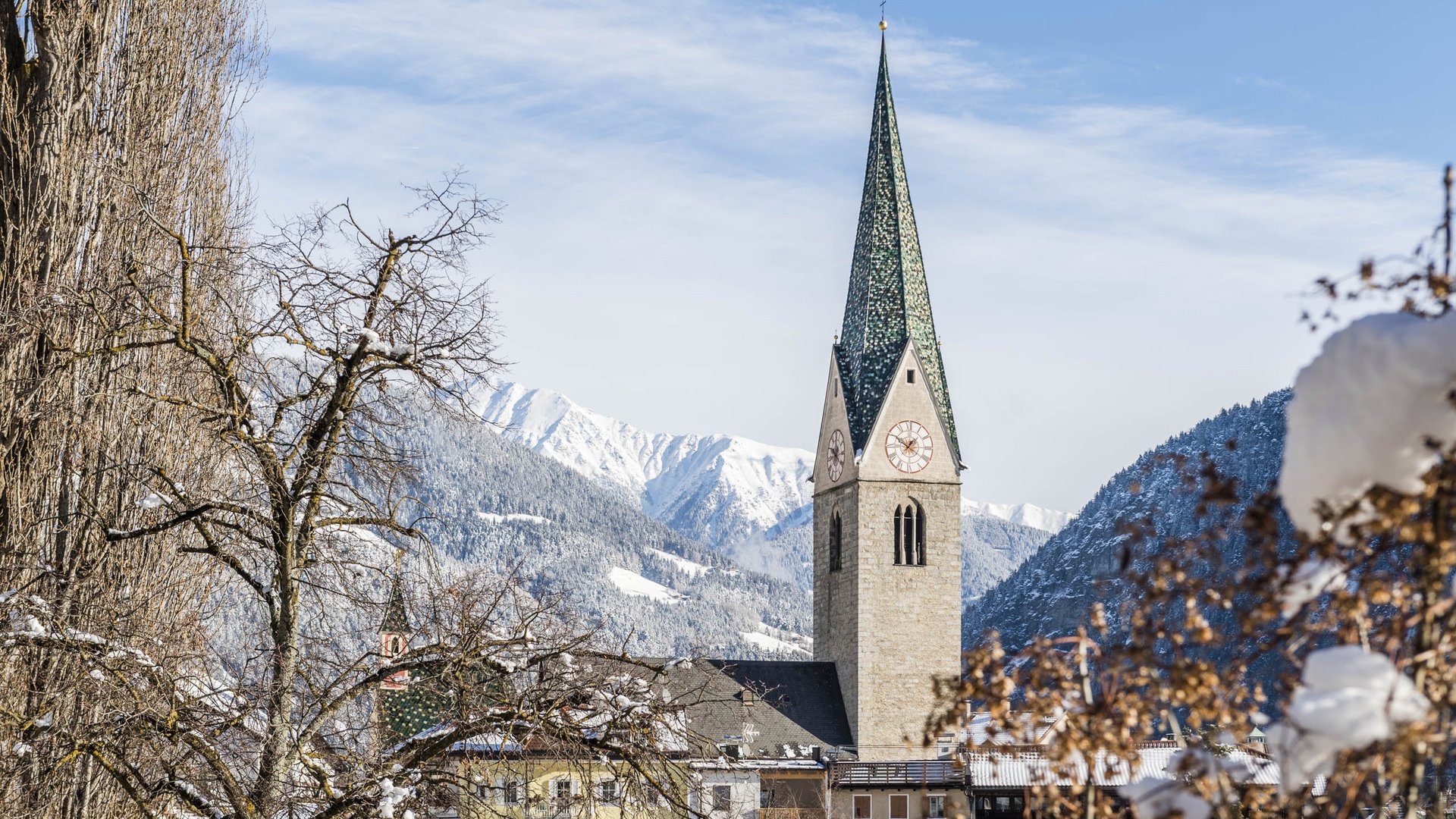 Mühlbach in the Pustertal valley The picture shows a church with a tall green church tower in a snowy village. In the background, snow-covered mountains and a blue sky are visible, while in the foreground bare trees and bushes are covered with snow.