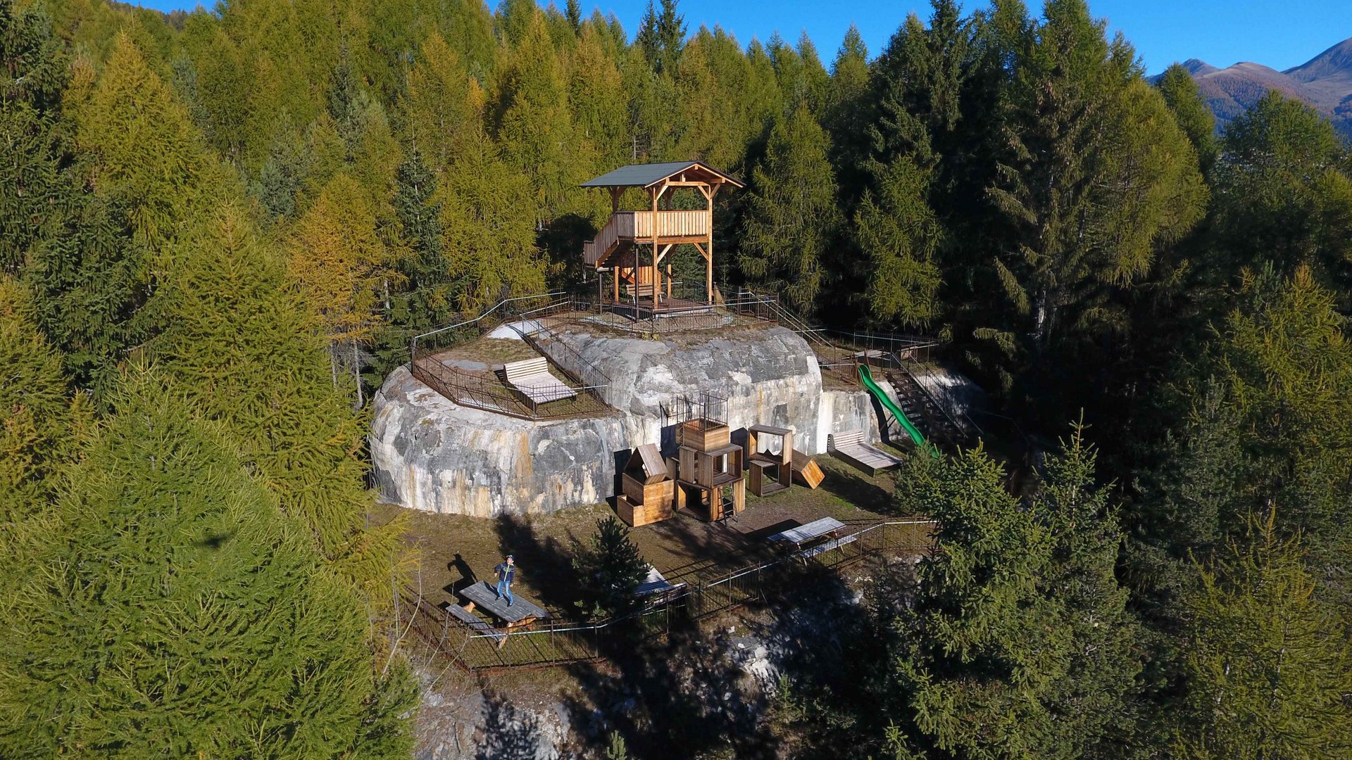 Het recreatiegebied in Spinges/Zuid-Tirol De afbeelding toont een rotsachtige heuvel in een bosgebied, waarop een houten constructie zoals een uitkijkpunt of een platform is gebouwd. Op de voorgrond bevinden zich zitplaatsen, speeltoestellen en een glijtoren, omringd door dichte groene bomen en een heldere, blauwe lucht op de achtergrond.