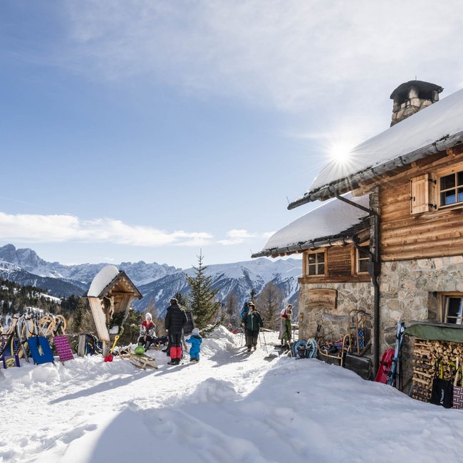 Kreuzwiesenalm A mountain hut stands in a wintery landscape with snow-covered mountains in the background. In front of the hut are numerous sleds and several people.