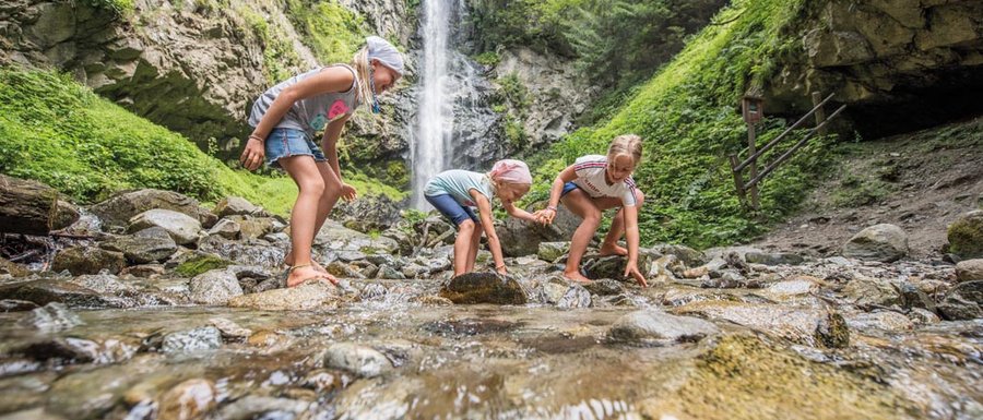 Find your accommodation in South Tyrol The picture shows three children playing barefoot in a shallow stream. In the background, there is a large waterfall cascading down a steep rock face, surrounded by lush, green forest.