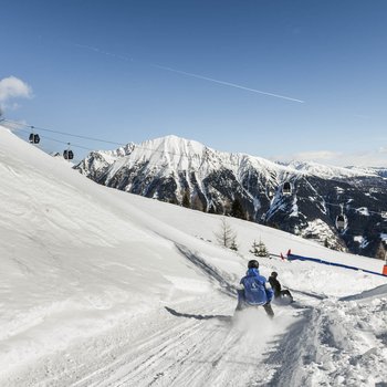 Willkommen in der Ferienregion Gitschberg Jochtal! Personen rodeln auf verschneiter Piste mit Seilbahn und Bergen im Hintergrund