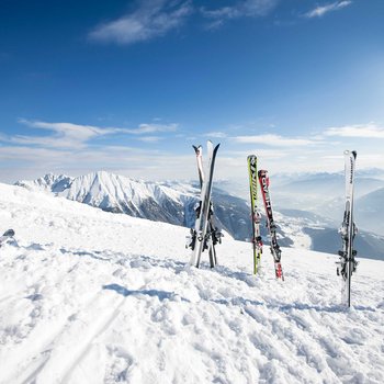 Willkommen in der Ferienregion Gitschberg Jochtal! Mehrere Skier stehen im Schnee vor schneebedeckten Bergen unter blauem Himmel