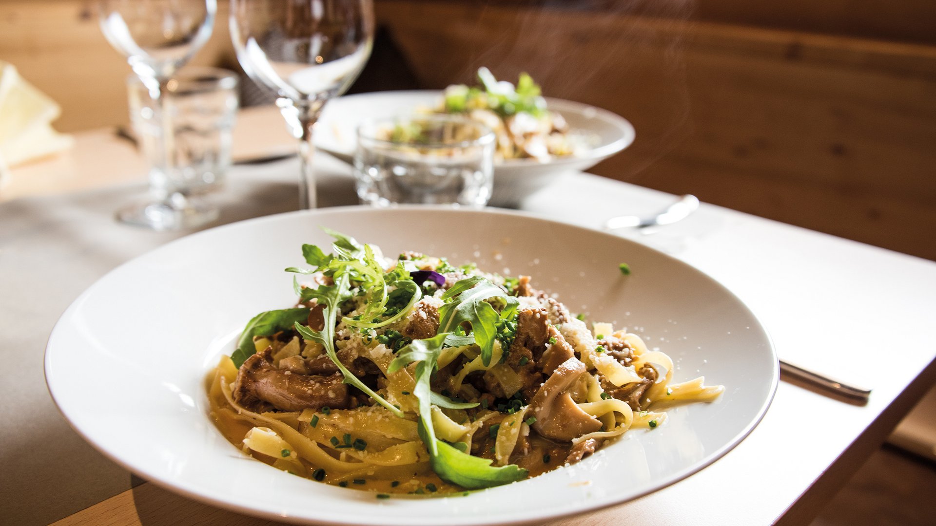 Discover the Alpine huts in Meransen, Vals & Co. The picture shows a finely prepared dish of pasta, garnished with mushrooms and fresh green herbs, served on a white plate in a cozy restaurant with wine glasses and other dishes in the background.