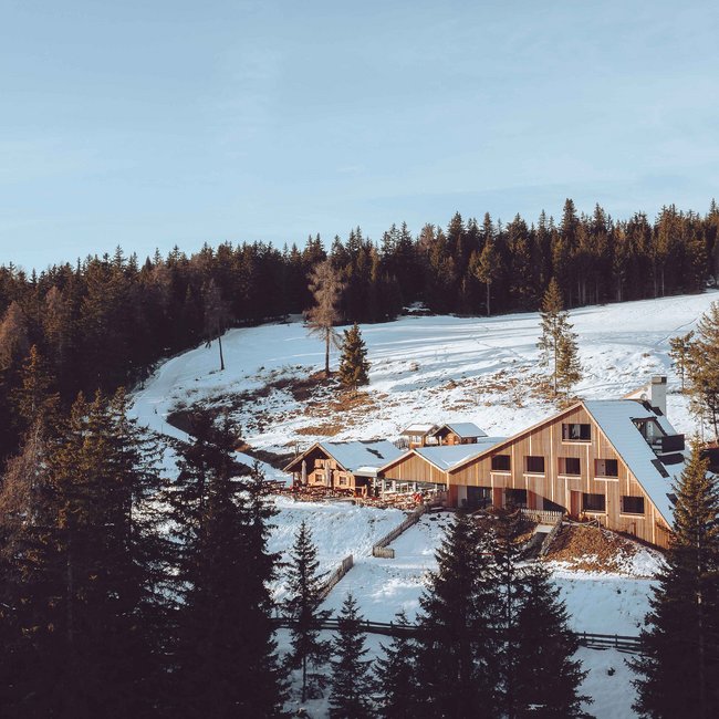 Oberhauserhütte De afbeelding toont een moderne houten berghut, omringd door met sneeuw bedekte heuvels en dicht bos. De hut ligt in een schilderachtig winterlandschap met dennenbomen op de voorgrond en een heldere, blauwe lucht erboven.