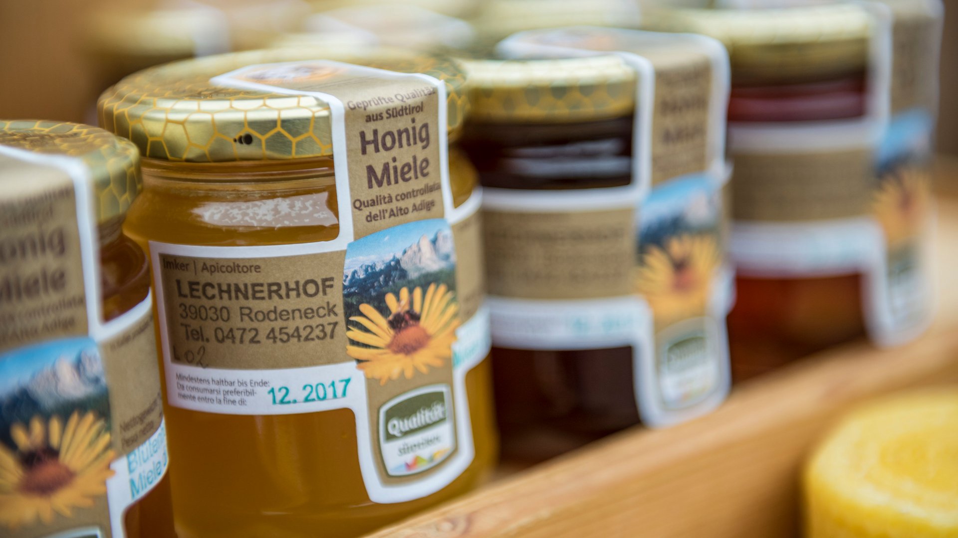 Nakupování v Mühlbachu/Itálie Obrázek ukazuje několik sklenic medu stojících na polici. Na štítcích sklenic je napsáno "Honig Miele" spolu s adresou a kontaktními údaji včelaře, který působí na statku nazvaném "Lechnerhof" v Rodenecku v Jižním Tyrolsku. Sklenice mají zlaté víčka s plástvovým vzorem.