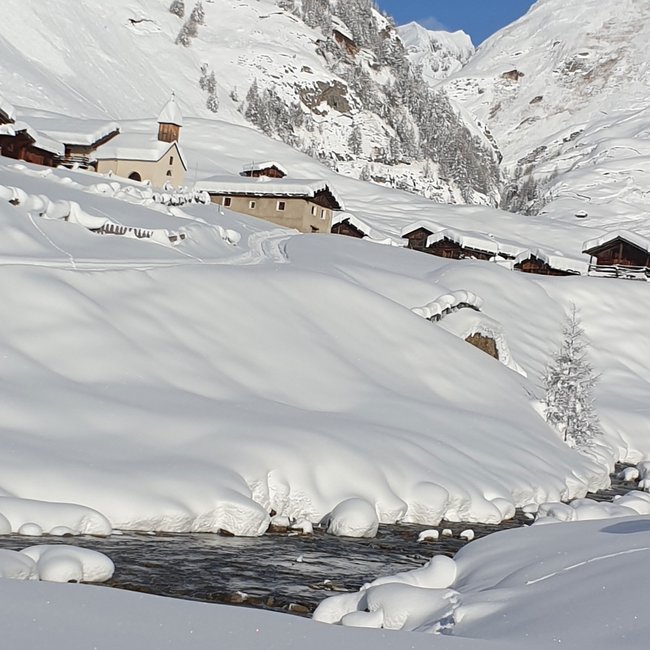 Zingerle Hütte Obrázek zobrazuje hluboce zasněženou horskou krajinu s malou skupinkou chat a kostelem v údolí. V popředí protéká malý potok zasněženou krajinou a v pozadí jsou vysoké zasněžené hory pod jasnou modrou oblohou.