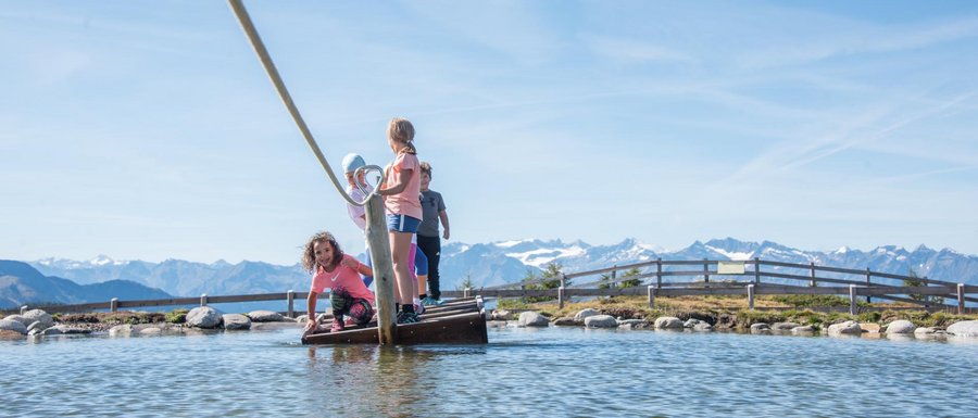 Find your accommodation in South Tyrol The picture shows several children standing on a small raft, which is being pulled with a rope across a shallow pond. In the background, there are mountains that complement the clear blue sky and the picturesque landscape.