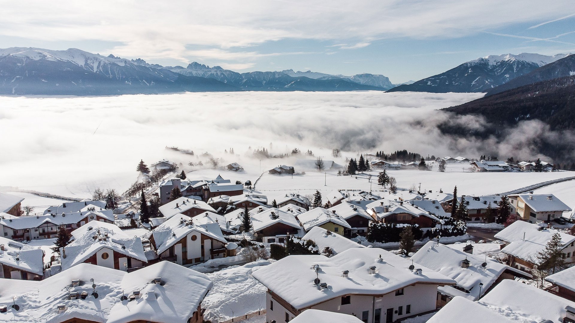 Das Bergdorf Meransen Das Bild zeigt ein Dorf mit schneebedeckten Häusern auf einem Hügel, das von einer Nebeldecke umgeben ist. Im Hintergrund sind beeindruckende, schneebedeckte Berggipfel unter einem teilweise bewölkten Himmel zu sehen.
