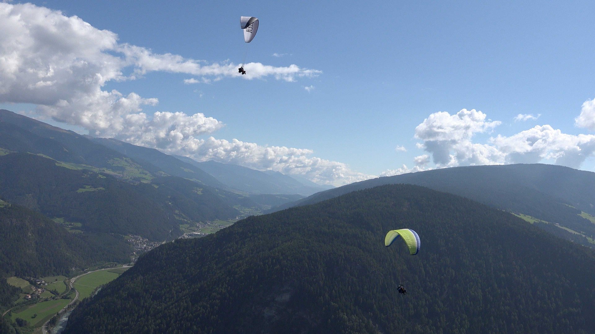 Paragliden in Zuid-Tirol De afbeelding toont twee paragliders die over een uitgestrekt berglandschap met dicht beboste heuvels vliegen. De lucht is helder met enkele verspreide wolken, en op de achtergrond strekken groene valleien en meer bergen zich uit.