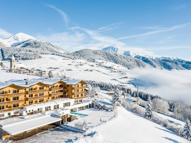 Willkommen in der Ferienregion Gitschberg Jochtal! Das Bild zeigt ein großes Hotel in einer schneebedeckten Winterlandschaft. Im Hintergrund sind schneebedeckte Berge, Wälder und eine kleine Kirche zu sehen, während das Hotel von strahlendem Sonnenschein erleuchtet wird.