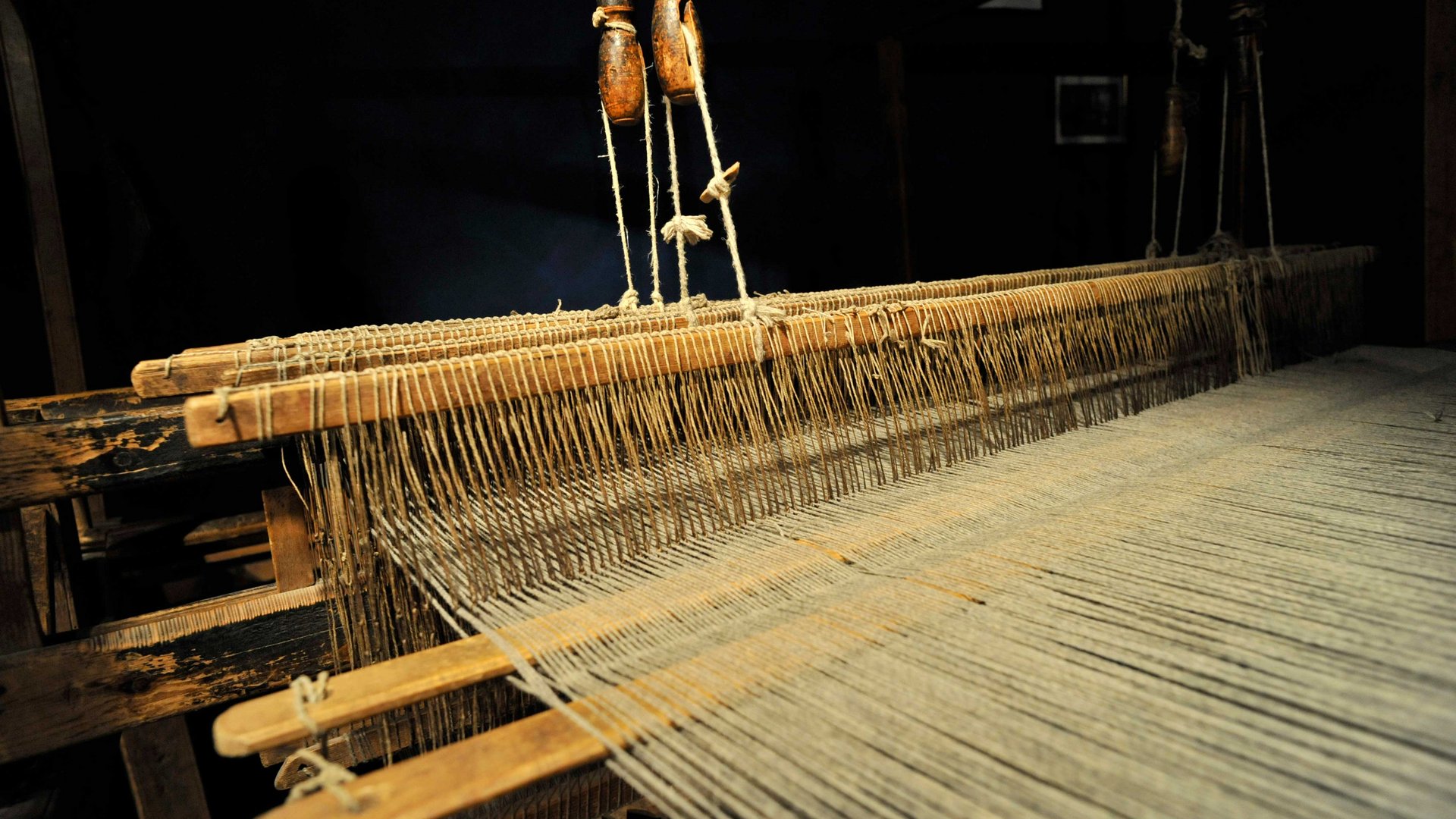 Zážitkový svět lodenu ve Vintlu/Pustertal Obrázek ukazuje starý dřevěný stav s nitěmi napnutými těsně vedle sebe. Stav je vystaven v tmavém prostředí, což umožňuje dobře vidět detaily mechaniky a nití.