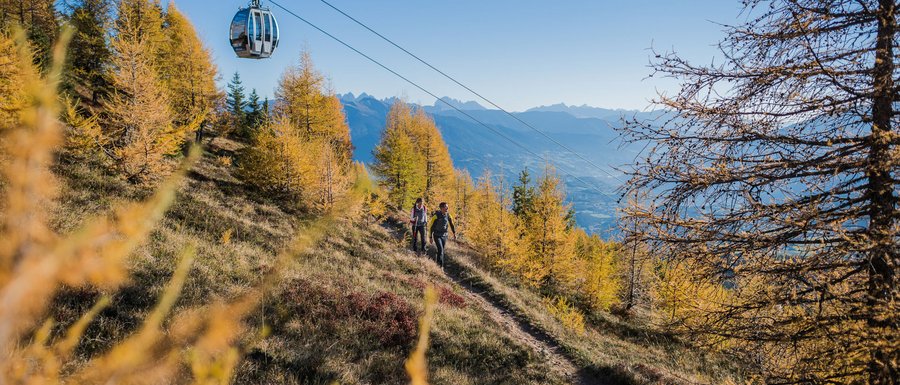 Find your accommodation in South Tyrol The picture shows a cable car traveling over a path used by hikers. The landscape is autumnal, with golden trees, and in the background, mountains can be seen under a clear blue sky.