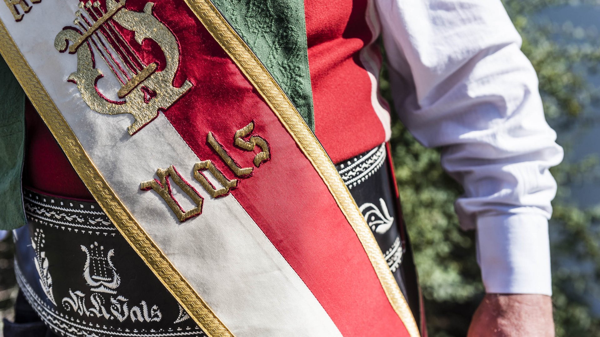 Vals: das Almendorf Das Bild zeigt den Ausschnitt einer traditionellen Tracht. Zu sehen ist eine rot-weiß-goldene Schärpe mit der Aufschrift „Vals“ und einem gestickten Emblem einer goldenen Harfe. Der Hintergrund ist unscharf, und der Fokus liegt auf der Kleidung und der Schärpe.
